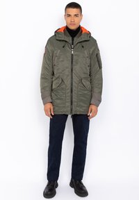 Zielona, ocieplana parka z pomarańczową podszewką kaptura, z zamkiem z przodu, kieszeniami po bokach oraz kieszenią na rękawie z zamkiem. Noszona z ciemnymi dżinsami i czarnymi botkami.