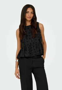 Femme aux longs cheveux foncés portant un top noir sans manches avec texture florale et un pantalon noir, debout avec une main dans la poche.