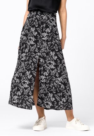 Femme portant une jupe midi à motifs floraux noirs et blancs avec une fente à l'avant et des baskets blanches, debout devant un fond uni.