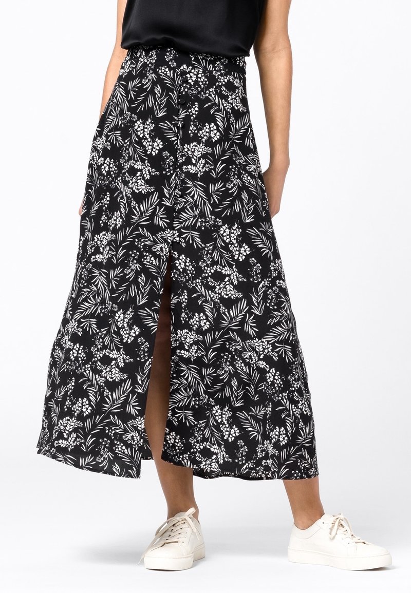 Femme portant une jupe midi à motifs floraux noirs et blancs avec une fente à l'avant et des baskets blanches, debout devant un fond uni.