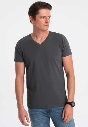 Camiseta de cuello en V de manga corta en gris oscuro, hecha de tela suave. El modelo la lleva con jeans de mezclilla azules y un reloj negro, posando frente a un fondo blanco.