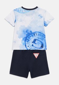 T-shirt blanc à manches courtes avec motif aquarelle bleu et logo circulaire, associé à un short bleu marine avec un patch logo triangulaire rouge.