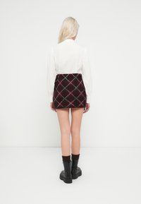 Γυναίκα με μακριά ξανθά μαλλιά, που φοράει λευκό πουκάμισο με μακριά μανίκια, μίνι φούστα μπορντό καρό, μαύρες μπότες και σκούρες κάλτσες, στέκεται με την πλάτη σε ένα λευκό τοίχο.