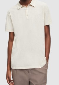 Polo de couleur crème avec patte à deux boutons et manches courtes. Texture douce et coupe décontractée, associé à un pantalon beige clair.