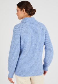 Suéter de punto azul claro con una textura acanalada, cuello alto y mangas largas. Presenta un ajuste ligeramente relajado y aberturas en los costados del dobladillo.