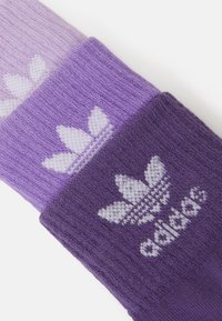 Három pár lila zokni eltérő árnyalatokban, kötött textúrával és fehér Adidas logóval, amely levélmintával díszített a mandzsettán.