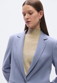 Ανοιχτό μπλε σακάκι με λεία υφή, δομημένους ώμους και κλείσιμο με ένα κουμπί, φορεμένο πάνω από μπεζ ριμπ μπλουζάκι με ζιβάγκο.