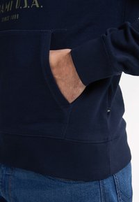 Felpa in cotone blu navy con tasca frontale, polsini e orlo a coste. Presenta dettagli di cucitura e un logo stampato sul petto.