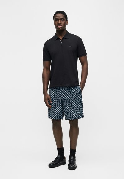 Άνδρας που στέκεται φορώντας μαύρο πόλο μπλουζάκι με κοντά μανίκια, μπλε σορτς με σχέδια, μαύρες κάλτσες και μαύρα αθλητικά παπούτσια μπροστά σε απλό φόντο.