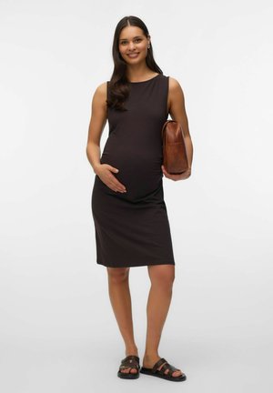 Femme enceinte en robe noire sans manches, tenant un sac marron sous un bras et reposant la main sur son ventre, portant des sandales noires.