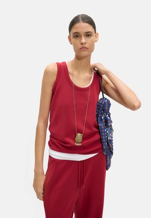 Młoda kobieta w czerwonym topie bez rękawów i spodniach, z długim złotym naszyjnikiem z zawieszką, trzymająca na ramieniu niebieski szal w wzory.