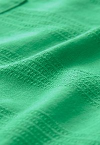 Grüner strukturierteter Stoff mit einem gewebten Muster, das subtile geometrische Designs aufweist. Das Material wirkt weich und leicht.