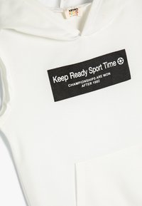 Vit huva sweatshirt med en svart rektangulär grafik med vit text och en ikon av en fotboll. Inkluderar en framficka.