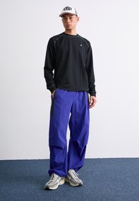Camisa negra de manga larga con escote redondo, pantalones morados de corte holgado con bolsillos laterales y zapatillas beige. Gorra de béisbol con logo.
