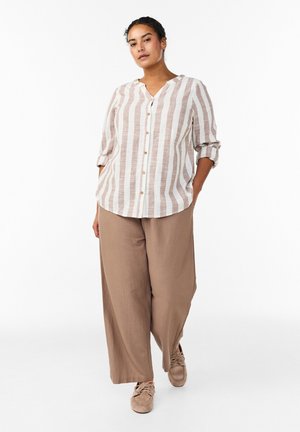 Žena oblečená v béžových širokých nohaviciach, béžovo-bielej vertikálne pruhovanej košeli s gombíkmi a vyhrnutými rukávmi, a béžových topánkach, stojaca s rukami v vreckách.