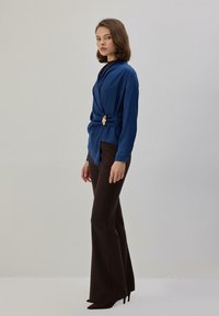 Top azul cruzado con un acento dorado en forma de hoja en la cintura, combinado con pantalones marrones acampanados de talle alto y tacones puntiagudos. Textura suave.