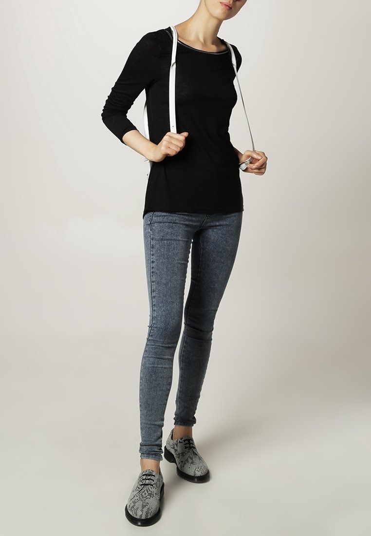 Czarna bluzka z długim rękawem, jasnoszare skinny jeans i teksturowane czarno-szare buty w wzory ze sznurowadłami. Białe paski zwisają z ramion.