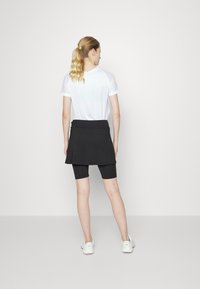 Camiseta atlética blanca con mangas de malla, combinada con una falda negra sobre pantalones cortos de ciclismo negros. El modelo lleva zapatillas blancas.