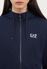 Marineblå hettegenser med glidelås laget av myk bomull, med snor i hetten og EA7-logo i hvitt på venstre bryst. Glidelåsdetalj synlig.