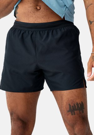 Mann trägt schwarze Sportshorts, zeigt Tätowierungen am rechten Oberschenkel und am linken Arm, hebt ein hellblaues Shirt an, um den Bauch freizulegen.