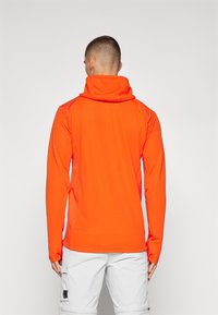 Orange hooded long-sleeve top with textured fabric and fitted design. Features a high collar and fitted cuffs, worn with white cargo pants.