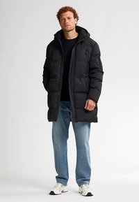 Sort polstret jakke med hætte, der har to forlommer foran og en lynlås. Kombineret med lyseblå jeans og hvide sneakers.