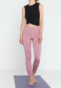 Mustaton hihaton paita, joka on solmittu vyötärölle, yhdistettynä istuviin vaaleanpunaisiin legginsseihin, joissa on tekstuurit yksityiskohtina nilkoissa. Lattiamatto alapuolella.