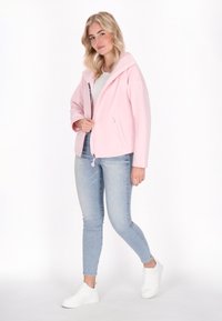 Mlada ženska v svetlo roza jakni, belem srajcu in modrih kavbojkah samozavestno pozira pred belo ozadjem ter nosi bele superge.