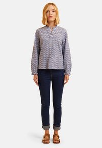 Blusa blu con motivo floreale, dal collo rotondo, con chiusura a bottoni e maniche lunghe, abbinata a jeans scuri e sandali marroni.