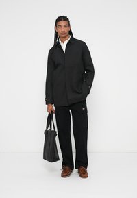 Cappotto leggero nero con colletto, chiusura a bottoni e tasche laterali. Abbinato a pantaloni cargo neri e scarpe da barca marroni, mentre tiene una borsa a tracolla nera.