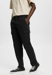 Svarta cargo-byxor i bomull med sidofickor, en avslappnad passform och en klassisk design. Kommer med svarta sneakers och en beige t-shirt.