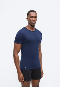 Άνδρας που στέκεται φορώντας μπλε στρατιωτικό στενό κοντομάνικο πουκάμισο και μαύρο σορτς, κοιτάζοντας ελαφρώς προς τα αριστερά με ουδέτερη έκφραση.