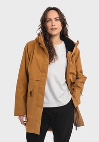 Braune wasserdichte Jacke mit Kapuze, Reißverschluss, zwei Seitentaschen und einer strukturierten Oberfläche, getragen über einem weißen Hemd und schwarzen Hosen.