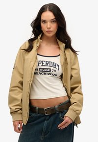 Veste bomber beige avec une fermeture éclair, des poignets côtelés et une texture lisse, portée par-dessus un débardeur blanc avec du texte bleu marine. Jupe en denim avec une ceinture.