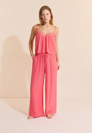 Junge Frau steht barfuß und trägt ein lockeres pinkfarbenes Pyjama-Set mit Spaghettiträger-Oberteil und weit geschnittenen Hosen vor einem schlichten beigen Hintergrund.