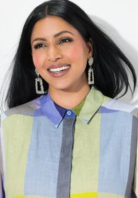 Camicia di flanella multicolore nelle tonalità di verde, blu e grigio, con chiusura a bottone blu e colletto. Orecchini a goccia geometrici brillanti.