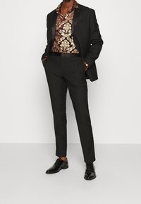 Černý oblek s lesklým saténovým klopem, doplněný vzorovanou košilí v zlatých a vínových tónech. Černé lesklé boty dokončují vzhled.