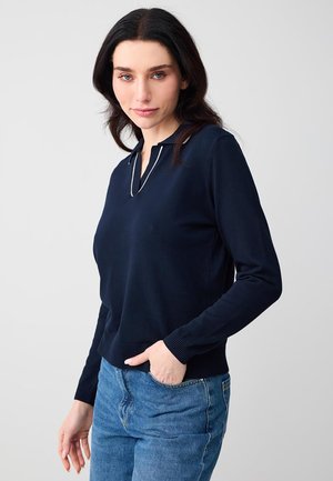 Femme aux cheveux bruns, portant un haut à manches longues bleu marine avec des bordures blanches et un jean bleu, debout avec une main dans la poche devant un fond uni.