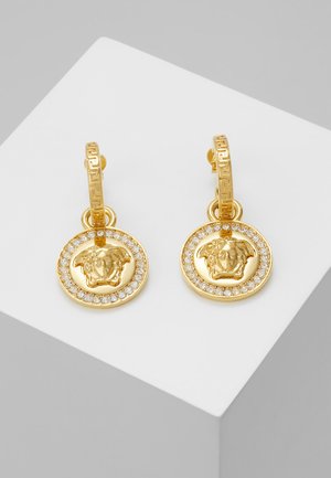 Les boucles d'oreilles en or présentent un design circulaire avec un motif de lion au centre, entouré d'une bordure de petites cristaux transparents. Créoles en or texturées.