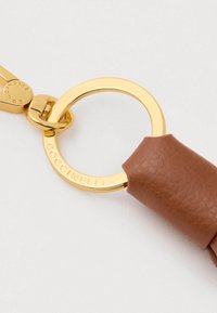 Inel de aur gravat cu „Coccinelle” atașat de o curea din piele maro pe un fundal alb.