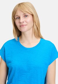 Jasnoniebieska koszulka z krótkim rękawem z teksturowanym geometrycznym wzorem i okrągłym dekoltem. Miękka tkanina, luźny krój i minimalne zdobienia.