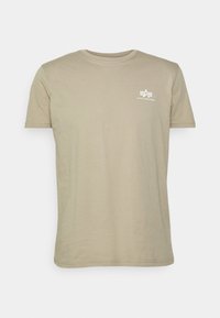 Beige tričko s krátkým rukávem a kulatým výstřihem se malým bílým logem Alpha Industries na levé straně hrudi.