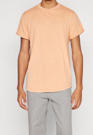 Mężczyzna w brzoskwiniowej koszulce z krótkim rękawem i dekoltem typu crew neck oraz jasnoszarych spodniach, stojący z ramionami swobodnie opuszczonymi na tle jednolitego tła.