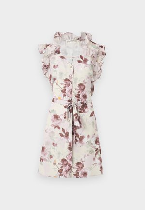 Donna in piedi che indossa un vestito senza maniche a stampa floreale, lunghezza al ginocchio, con spalle arricciate e tacchi marroni a punta su sfondo bianco.