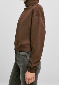 Persona que lleva un suéter de cuello alto marrón y unos vaqueros negros desgastados, vista de perfil contra un fondo claro y liso.