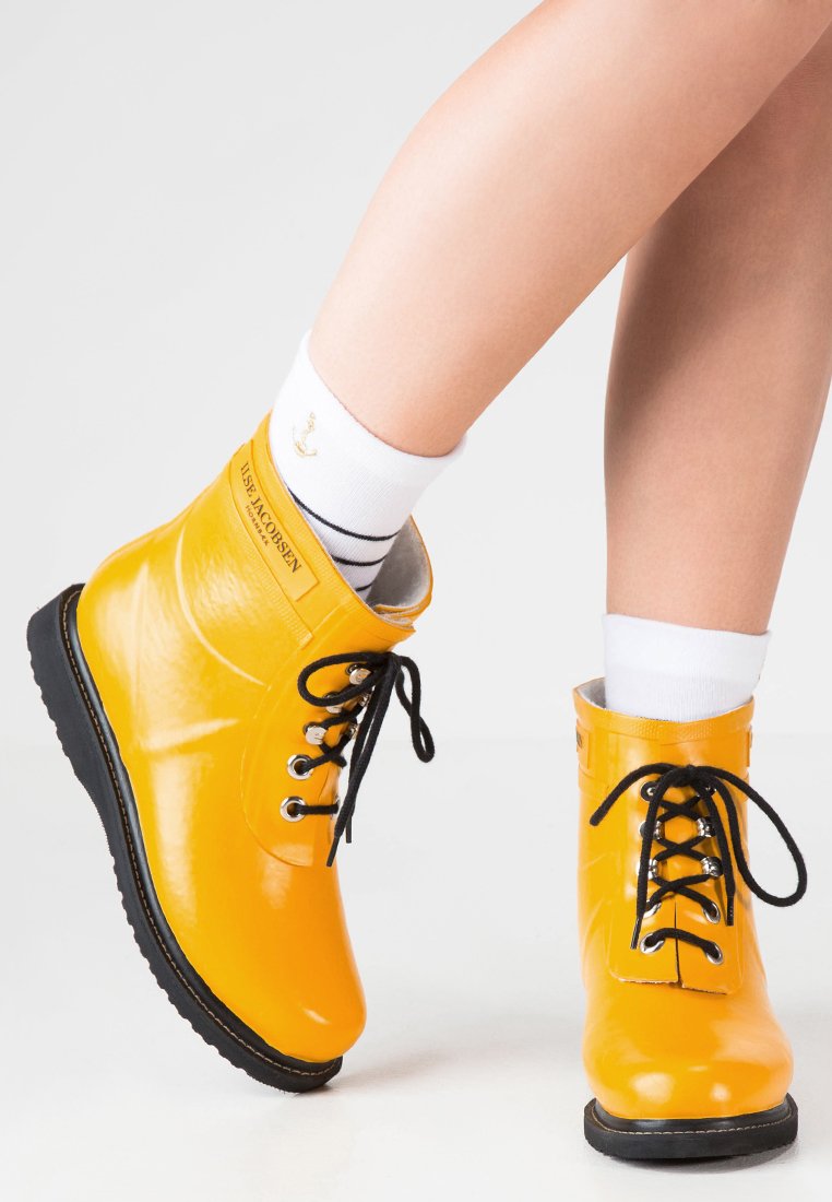 Geel rubberen enkelboots met zwarte veters en zool, voorzien van een gladde glanzende afwerking. Een merkdetail is zichtbaar aan de zijkant.