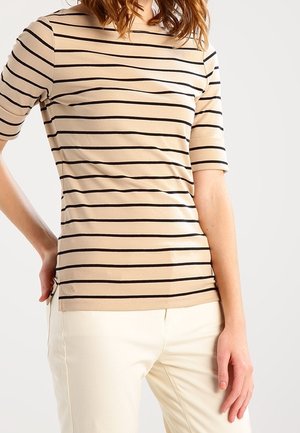 Vrouw draagt een beige top met korte mouwen met zwarte horizontale strepen en offwhite broek, staand met één hand op de heup tegen een effen achtergrond.