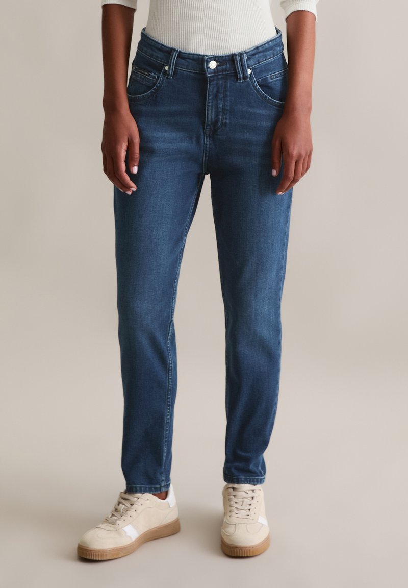 Denim jeans in een medium blauwe wassing, met een smalle taille, rechte pijpen en een klassiek vijf-pocket ontwerp. Gecombineerd met beige sneakers.