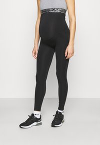 Leggings de maternidade pretos com uma faixa larga e estampada. A modelo combina-os com sapatilhas pretas e meias brancas até ao tornozelo.