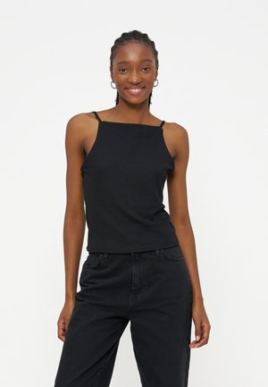 Γυναίκα που χαμογελά με πλεγμένα μαλλιά, φοράει σκουλαρίκια σε σχήμα κρίκου, μαύρο αμάνικο τοπ με λεπτές τιράντες και μαύρο παντελόνι, μπροστά σε λευκό φόντο.